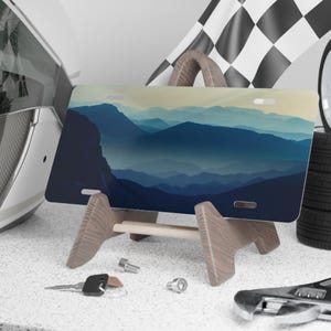 K&ouml;nnte beinhalten: Ein Nummernschild mit einem Berglandschaftsdesign in Blau- und Gr&uuml;nt&ouml;nen, pr&auml;sentiert auf einem Holzst&auml;nder. Die Platte wird mit Rennsport-Accessoires wie einer Zielflagge, Reifen und einem Helm ausgestellt.