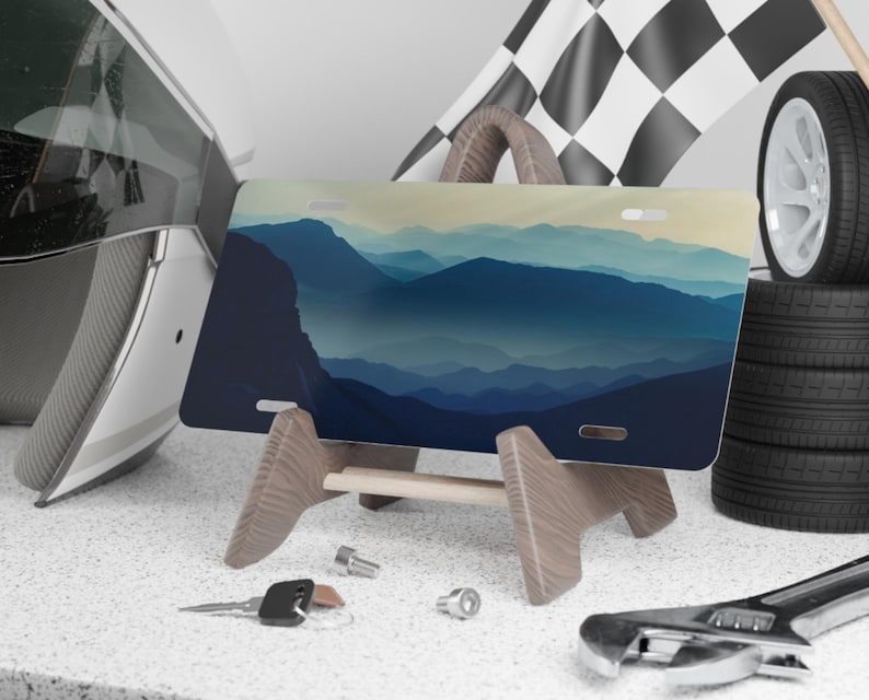 K&ouml;nnte beinhalten: Ein Nummernschild mit einem Berglandschaftsdesign in Blau- und Gr&uuml;nt&ouml;nen, pr&auml;sentiert auf einem Holzst&auml;nder. Die Platte wird mit Rennsport-Accessoires wie einer Zielflagge, Reifen und einem Helm ausgestellt.