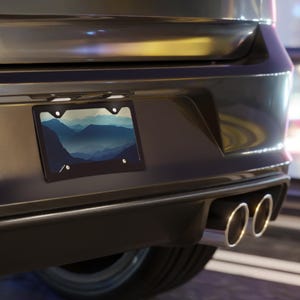 K&ouml;nnte beinhalten: Schwarzer Kennzeichenrahmen mit einem Berglandschaftsdesign. Das Kennzeichen zeigt ein blau-wei&szlig;es Gradientenbild von Bergketten. Der Rahmen ist an der R&uuml;ckseite eines dunkelgrauen Fahrzeugs angebracht.