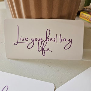 Puede incluir: Tarjeta blanca con el texto "Live your best tiny life." escrito en cursiva púrpura.
