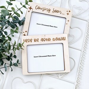 May include: Two wooden picture frames against a white background. The top frame reads "Coming Soon..." and the bottom frame reads "Here we grow again!" Both frames have a space for an ultrasound photo and are decorated with stars.