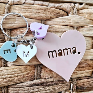 May include: A pink heart-shaped keyring with the word "mama" inscribed on it. The keyring has three smaller heart charms attached to it, one light blue with the letter "E", one white with the letter "M", and one purple with a small heart design.