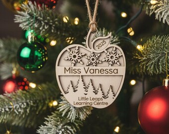 May include: Wooden apple-shaped Christmas ornament with laser-cut snowflakes, trees, and the text "Miss Vanessa" and "Little Leaps Learning Centre". The ornament is hanging on a Christmas tree with red and gold ornaments.