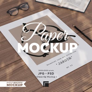 May include: A paper mockup with the word "Paper MOCKUP" in large white letters. The design features a beige square with black brushstrokes and the text "Botanical Line Art Collection 'JARDIN' NO 02". A pair of glasses, a pencil, and an open book are also visible.