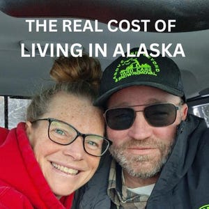 May include: A photograph of two people smiling, with text that reads "THE REAL COST OF LIVING IN ALASKA" and "$10,000's MORE Per Year." The man wears a black cap and sunglasses. The woman wears glasses and a red hoodie.