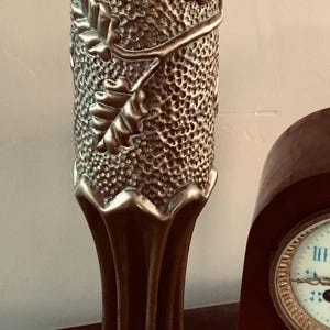 Puede incluir: Un jarrón alto, plateado, con una superficie texturizada y un diseño de hojas de roble en relieve. El jarrón tiene una base acampanada y un cuello estrecho. Un reloj de madera es parcialmente visible en el fondo.