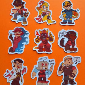 Op de afbeelding: Een verzameling van negen kleurrijke cartoonkarakterstickers op een oranje achtergrond. De stickers tonen verschillende personages in actieposes, met verschillende outfits en accessoires. De personages zijn in verschillende kleuren, waaronder rood, geel, blauw en bruin.