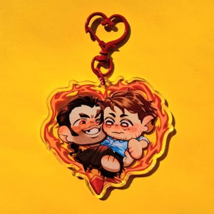 Puede incluir: Un llavero en forma de corazón con una ilustración de dibujos animados de dos figuras abrazándose, rodeadas por un borde de fuego. El llavero tiene un cierre rojo en forma de corazón. Las figuras tienen cabello oscuro y visten ropa azul y negra.