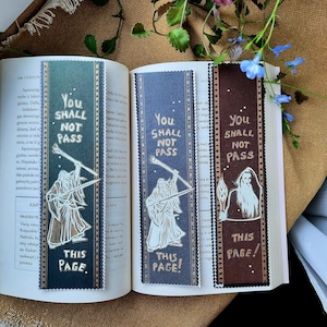Peut inclure: Trois marque-pages avec une silhouette blanche d'une figure tenant une épée et le texte "You Shall Not Pass" en lettres dorées. Les marque-pages sont sur un livre avec du texte dans une langue étrangère. Les marque-pages sont vert, bleu et marron.