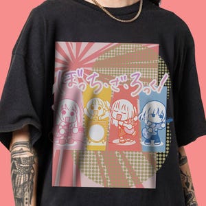 May include: Black t-shirt featuring a colorful graphic of four anime-style characters playing instruments. The design includes Japanese text and a pink, yellow, and blue color scheme. The shirt has a crew neck and short sleeves.