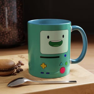 May include: A teal blue ceramic mug with a cartoon face and buttons on the front. The face is green and has a wide smile. The buttons are yellow, red, green, and blue.