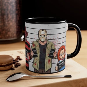 May include: A black and white ceramic mug featuring a horror movie theme. The mug displays illustrations of Freddy Krueger, Jason Voorhees, and Chucky, each holding a police lineup sign. The mug has a black interior and handle.