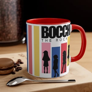 May include: A white ceramic mug with a red rim and handle. The mug features a colorful striped background with the text "BOCCI THE ROCK" in black. Silhouettes of four anime characters are depicted in front of the text.