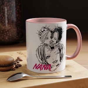 May include: White ceramic mug with a pink interior and handle. The mug features a black and white illustration of two anime characters in a heart shape, with the word "NANA" in pink.