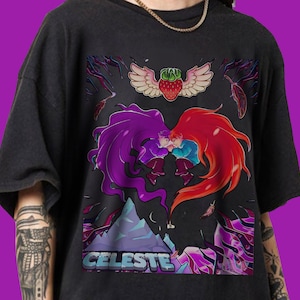 May include: Black t-shirt featuring a graphic from the video game Celeste. The design includes two figures with flowing hair, one purple and one red, embracing. A winged strawberry and the word "CELESTE" are also visible.