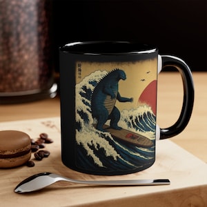 May include: Black ceramic mug featuring a blue Godzilla surfing a wave, with a red sun and Japanese text. The design is reminiscent of classic Japanese art. The mug has a white interior and a black handle.