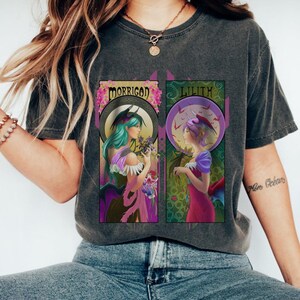 May include: Dark gray t-shirt featuring two Art Nouveau-style illustrations. One depicts a woman with teal hair labeled "Morrigad," the other a woman with purple hair labeled "Lilith." Both are set against a circular backdrop with floral and bat-like elements.