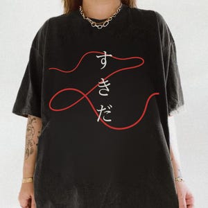 May include: A black short-sleeved t-shirt featuring a graphic of a continuous red line forming a loose, abstract shape. Centered within the red line are the white Japanese characters すきだ, arranged vertically. The shirt has a crew neckline.