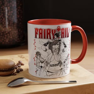 May include: White ceramic mug with a red interior and handle. The mug features the text "FAIRY TAIL" in red, along with a black and white anime illustration of a female character. The mug is on a wooden surface.
