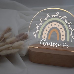 Op de afbeelding: Een houten basis met een helder acryl regenboog nachtlampje. De regenboog is in tinten van groen, oranje en beige met witte stippen. De naam "Clarissa" is in cursief geschreven met een hart ernaast.
