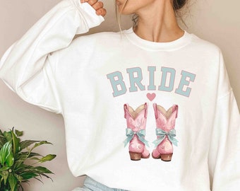 May include: White sweatshirt with the word "BRIDE" in blue text with a pink heart above it. The sweatshirt features a pink cowboy boot with a blue bow on each side.