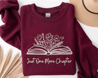 Peut inclure: Sweat-shirt bordeaux avec un graphique blanc d'un livre ouvert avec des fleurs qui en sortent. Le texte "Just One More Chapter" est imprimé sous le livre.
