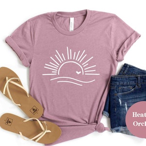 Pu&ograve; includere: Una t-shirt color orchidea heather con una grafica bianca di un sole con un cuore sopra un'onda. La t-shirt &egrave; stesa su uno sfondo bianco con un paio di sandali marroni e pantaloncini di jeans.