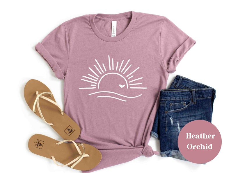 Pu&ograve; includere: Una t-shirt color orchidea heather con una grafica bianca di un sole con un cuore sopra un'onda. La t-shirt &egrave; stesa su uno sfondo bianco con un paio di sandali marroni e pantaloncini di jeans.