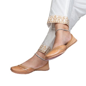 May include: A pair of tan leather shoes with decorative stitching and a pointed toe. The shoes are worn by a person wearing white pants with gold embroidery.