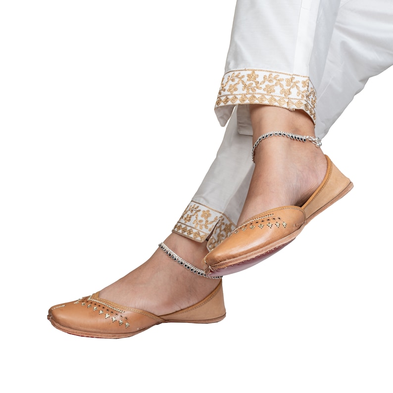 May include: A pair of tan leather shoes with decorative stitching and a pointed toe. The shoes are worn by a person wearing white pants with gold embroidery.