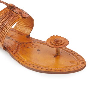 May include: Tan leather sandals with intricate floral designs and a braided strap. The sandals have a pointed toe and a small flower embellishment on the toe strap.