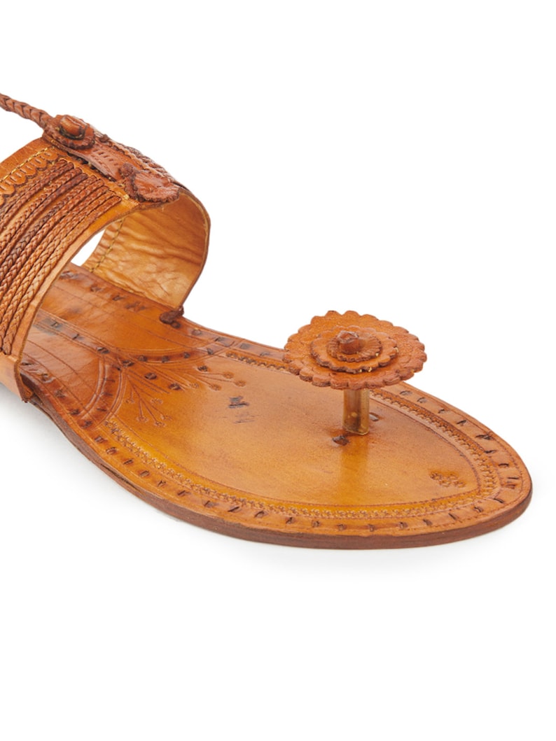 May include: Tan leather sandals with intricate floral designs and a braided strap. The sandals have a pointed toe and a small flower embellishment on the toe strap.