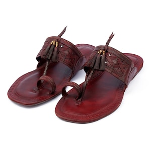 Könnte beinhalten: Ein Paar burgunderfarbene Ledersandalen mit aufwendigen Designs und Quasten. Die Sandalen haben eine Zehenschlaufe und einen Riemen, der um den Knöchel geht.