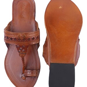 Pu&ograve; includere: Un paio di sandali in pelle marrone con intricati disegni traforati e un anello per le dita. I sandali hanno una superficie strutturata e una suola in gomma nera. I sandali sono esposti su sfondo bianco.