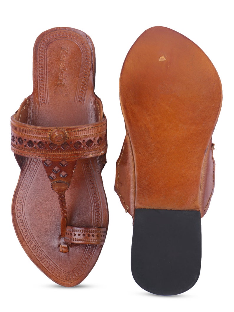 Pu&ograve; includere: Un paio di sandali in pelle marrone con intricati disegni traforati e un anello per le dita. I sandali hanno una superficie strutturata e una suola in gomma nera. I sandali sono esposti su sfondo bianco.