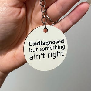 May include: A white, circular keyring with the black text "Undiagnosed but something ain't right". The keyring is attached to a silver chain and a silver ring.