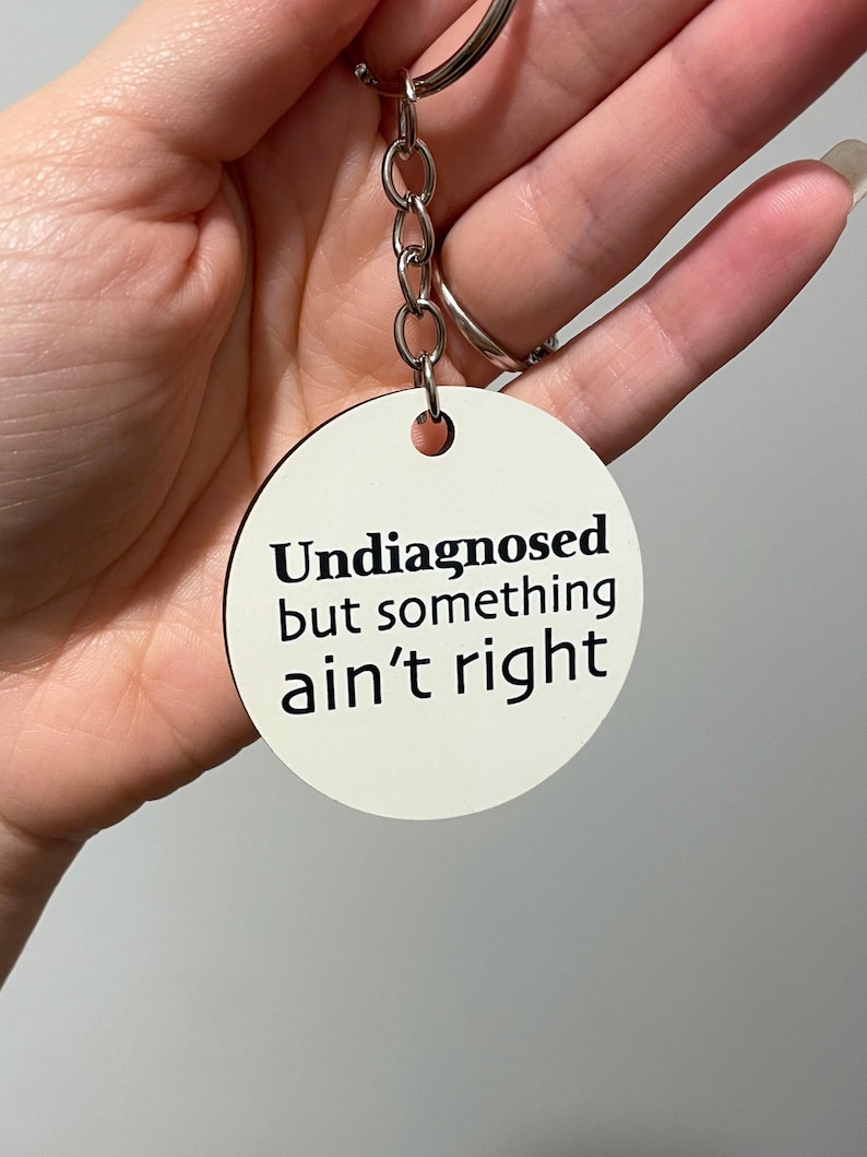 May include: A white, circular keyring with the black text "Undiagnosed but something ain't right". The keyring is attached to a silver chain and a silver ring.