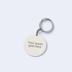 May include: A circular, cream-coloured keyring with a silver-coloured key ring and chain. The keyring features the text "Your quote goes here" in black. The background is a plain, light grey.