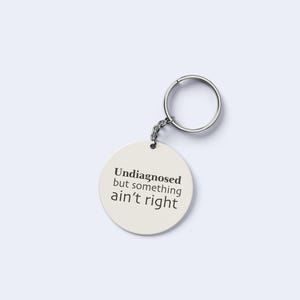 May include: An off-white circular keyring with the text "Undiagnosed but something ain't right" in black. It features a silver-coloured ring and chain.