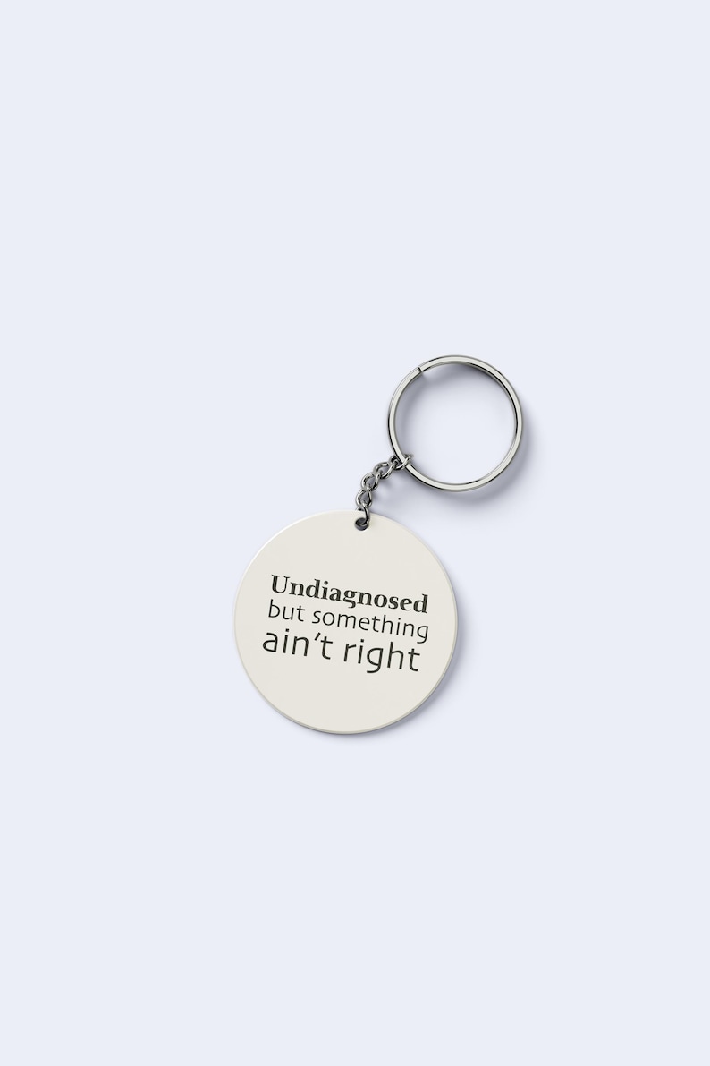 May include: An off-white circular keyring with the text "Undiagnosed but something ain't right" in black. It features a silver-coloured ring and chain.