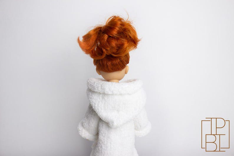 May include: A white hooded robe with a soft, fluffy texture. The robe is worn by a doll with long, red hair pulled back in a ponytail.