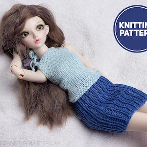 May include: A doll wearing a light blue knitted top and a dark blue knitted skirt. The doll has long brown hair and is lying on a white surface. A blue circle with the words "KNITTING PATTERN" is in the upper right corner.