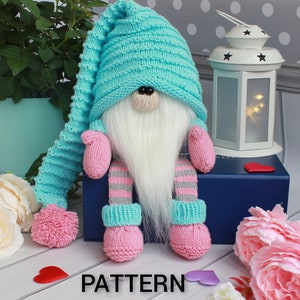 Puede incluir: Un gnomo de punto con un gorro azul, barba blanca y guantes rosas. El gnomo está sentado en una caja azul con una linterna blanca y flores rosas en el fondo. El texto "PATTERN" está impreso en la parte inferior de la imagen.