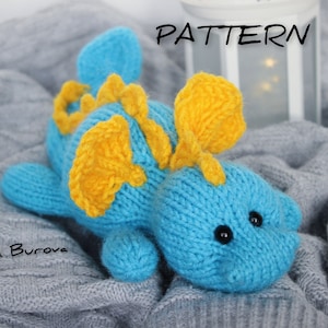May include: A blue knitted dragon toy with yellow wings and black eyes. The toy is lying on a grey knitted blanket. The word "PATTERN" is written in black on a white background.