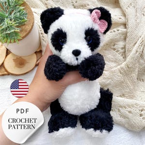 May include: A crochet pattern for a panda bear amigurumi toy. The panda is white with black ears, legs, and eye patches. It has a pink heart-shaped bow on its ear. The pattern is available as a PDF download.