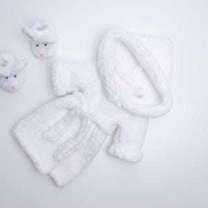 May include: White knitted baby robe with a hood and matching booties. The robe has a tie belt and is made of a soft, fluffy yarn.