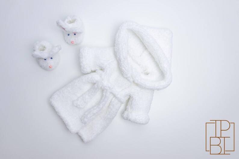 May include: White knitted baby robe with a hood and matching booties. The robe has a tie belt and is made of a soft, fluffy yarn.