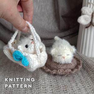 May include: Two small, fuzzy white crocheted kittens. One kitten rests inside a white crocheted basket with a bright blue bow, while the other kitten sleeps on a round brown crocheted cushion. The text KNITTING PATTERN is visible at the bottom.