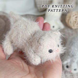 Puede incluir: Un pequeño gatito de punto de color beige claro, esponjoso, con ojos oscuros y nariz rosa. El texto PDF KNITTING PATTERN es visible en la parte superior. El gatito tiene un aspecto suave y texturizado, hecho de hilo con cabeza redondeada.