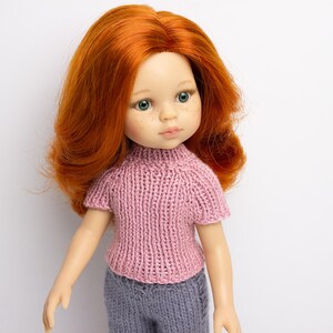 Pode incluir: Uma boneca com cabelo comprido e encaracolado vermelho usando um su&eacute;ter de tric&ocirc; rosa e cal&ccedil;as cinza. A boneca tem olhos verdes e uma tez clara.